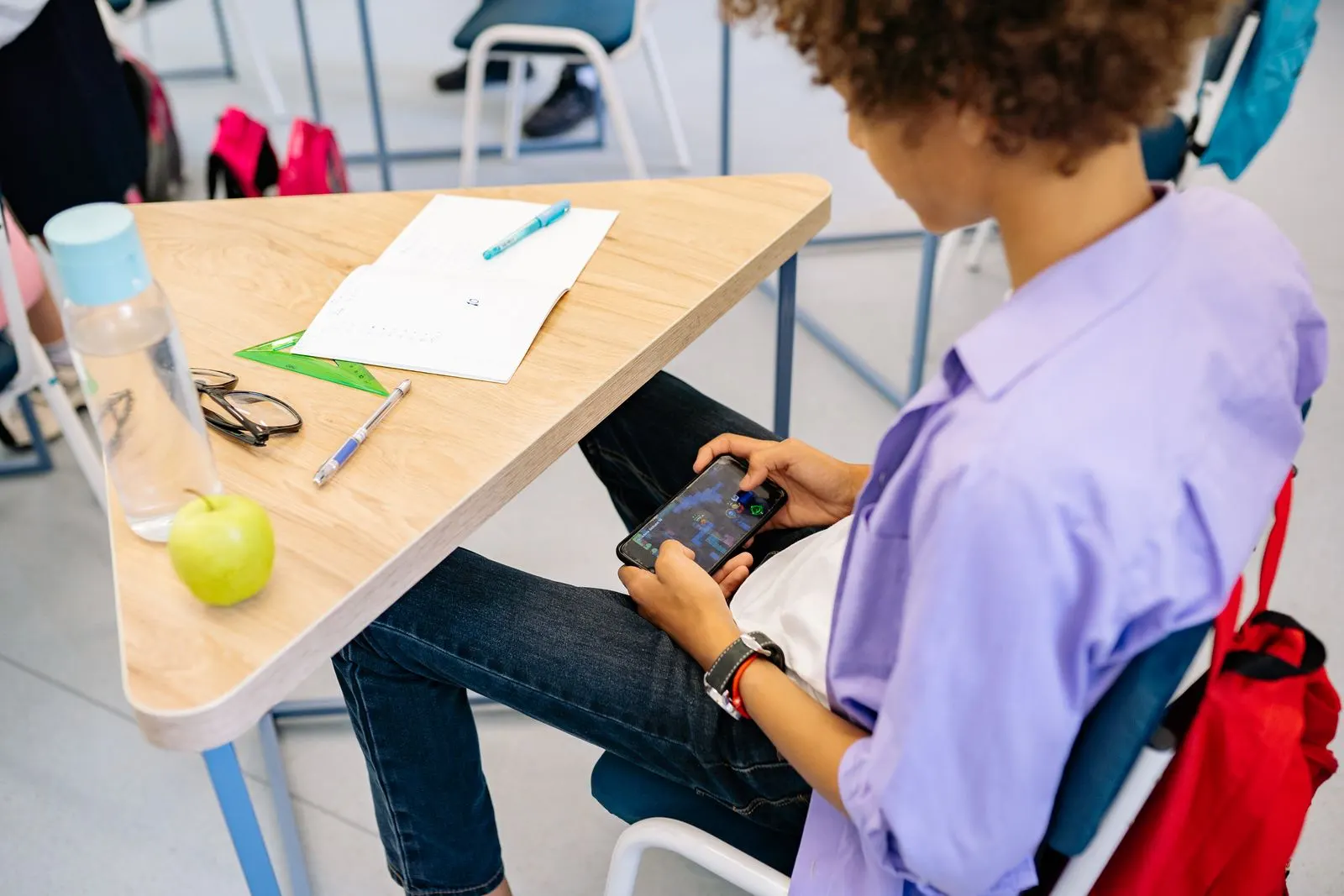 Ein Schüler spielt am Schulpult verstohlen ein Handyspiel unter dem Tisch, während Heft und Apfel unberührt vor ihm liegen
